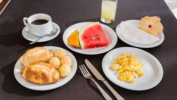 Una mesa de desayuno con platos de cruasanes, huevos revueltos, sandía, naranja y un vaso de jugo.
