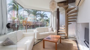Ein modernes Wohnzimmer mit einem weißen Sofa, einem Holztisch und einem großen Fenster mit Blick auf Palmen und Gebäude.