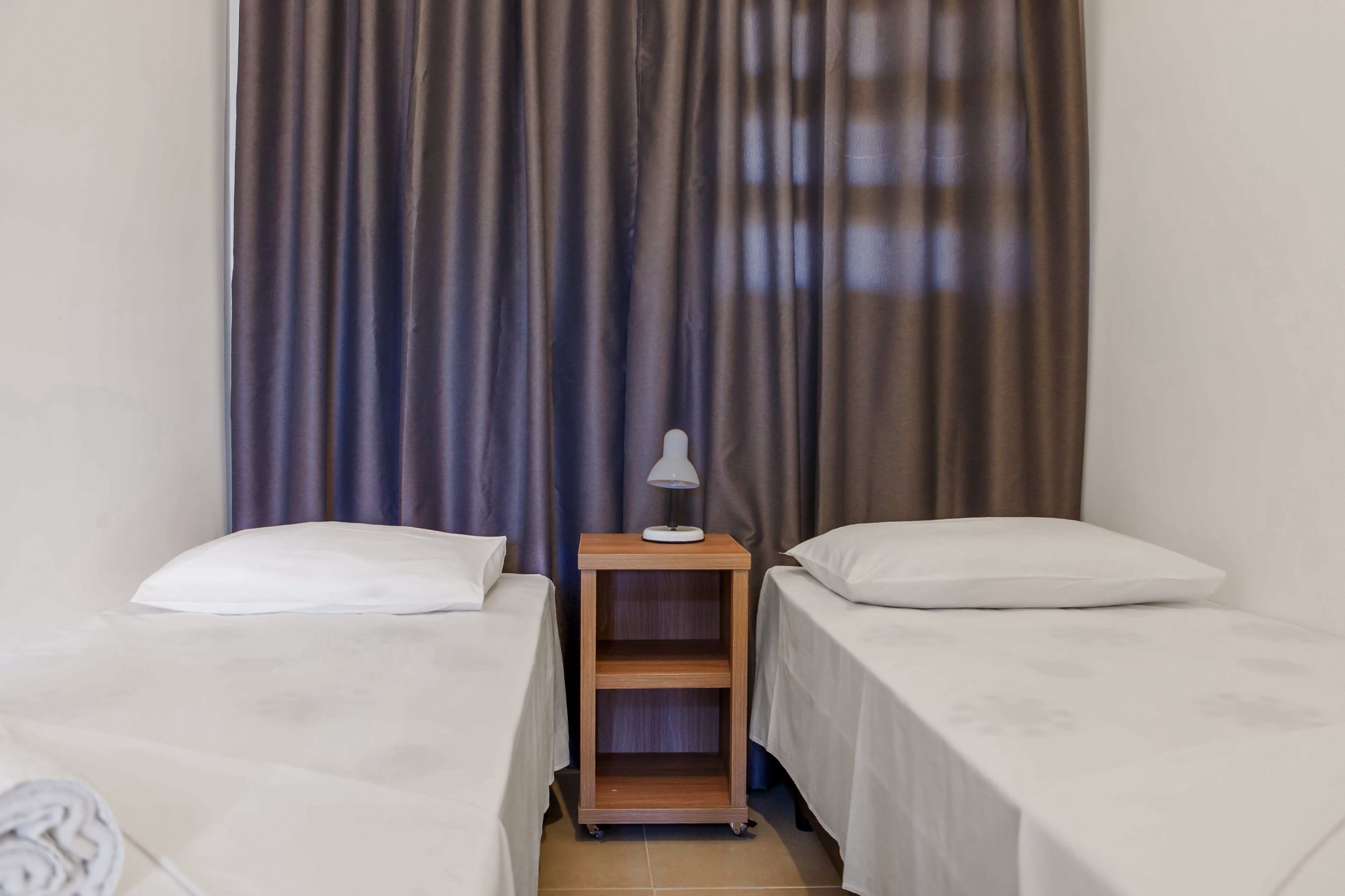 A hotel room with two single beds, a wooden nightstand with a lamp, and a curtain backdrop.