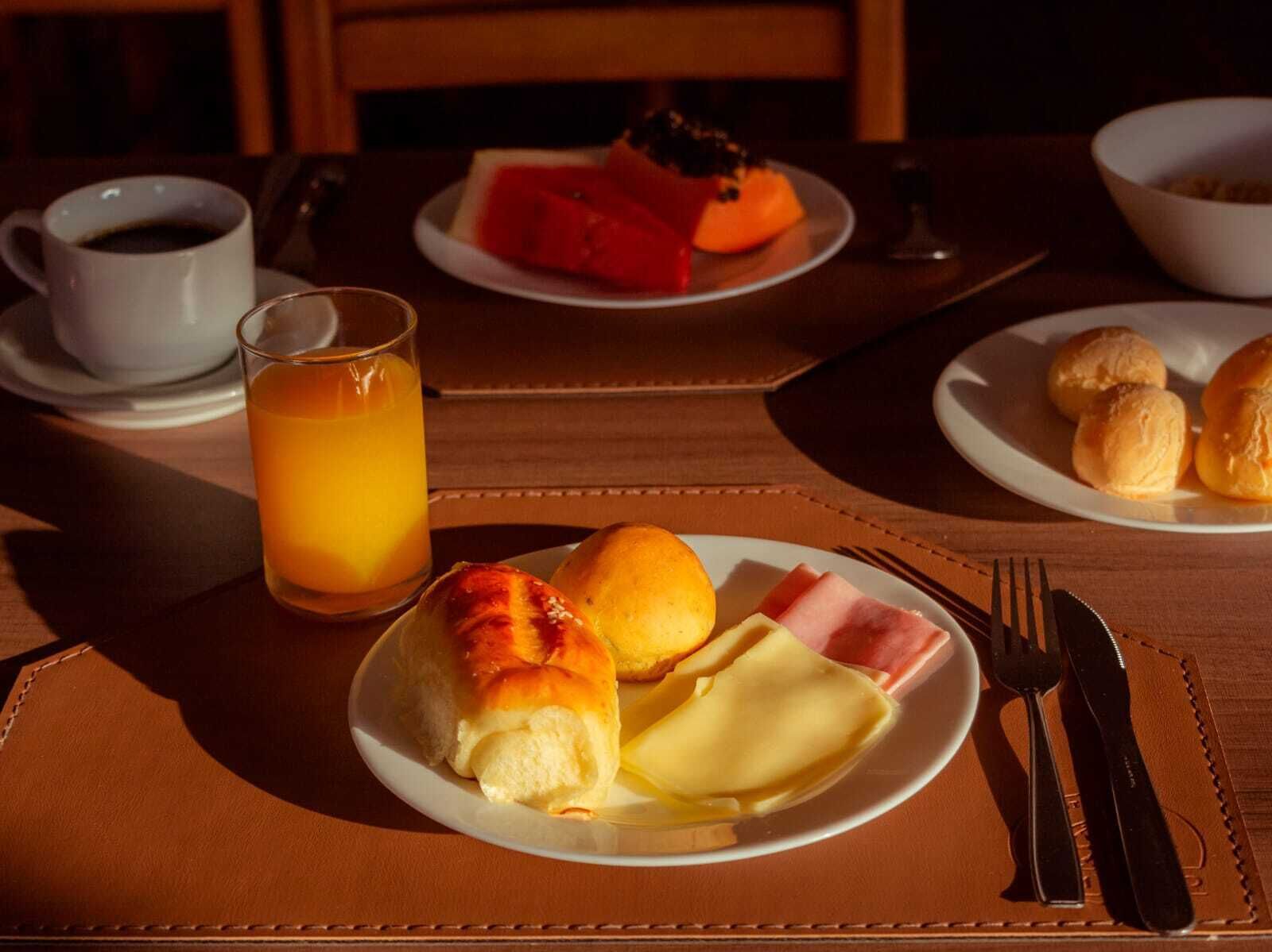 Un desayuno con un plato de panecillos, jamón y queso, un vaso de zumo de naranja y una taza de café sobre una mesa.