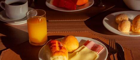 Un desayuno con un plato de panecillos, jamón y queso, un vaso de zumo de naranja y una taza de café sobre una mesa.