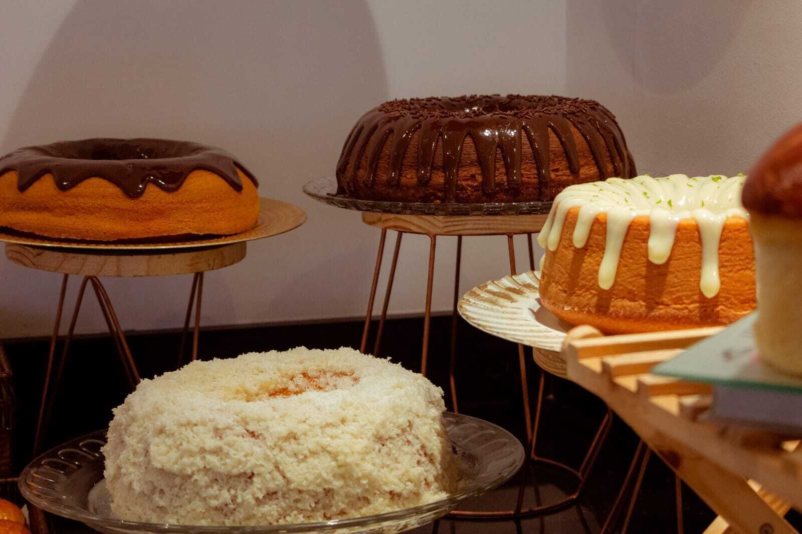A variety of cakes on display, including a bundt cake, a cake with white icing, and a cake with chocolate drizzle.