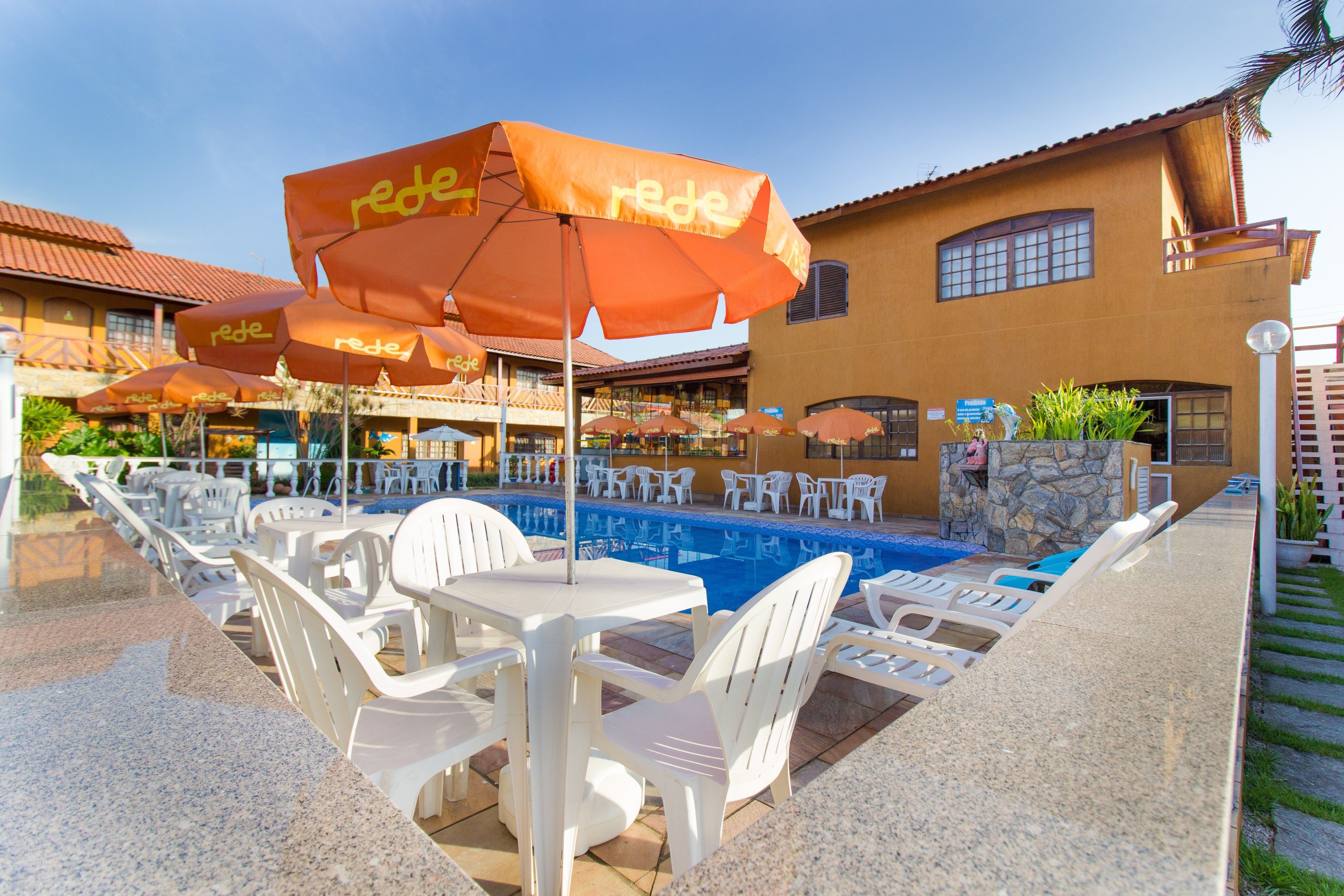 Un área junto a la piscina con sillas y mesas de plástico blanco, sombrillas naranjas con la palabra "rede" y un edificio con tejado de baldosas y detalles de piedra al fondo.
