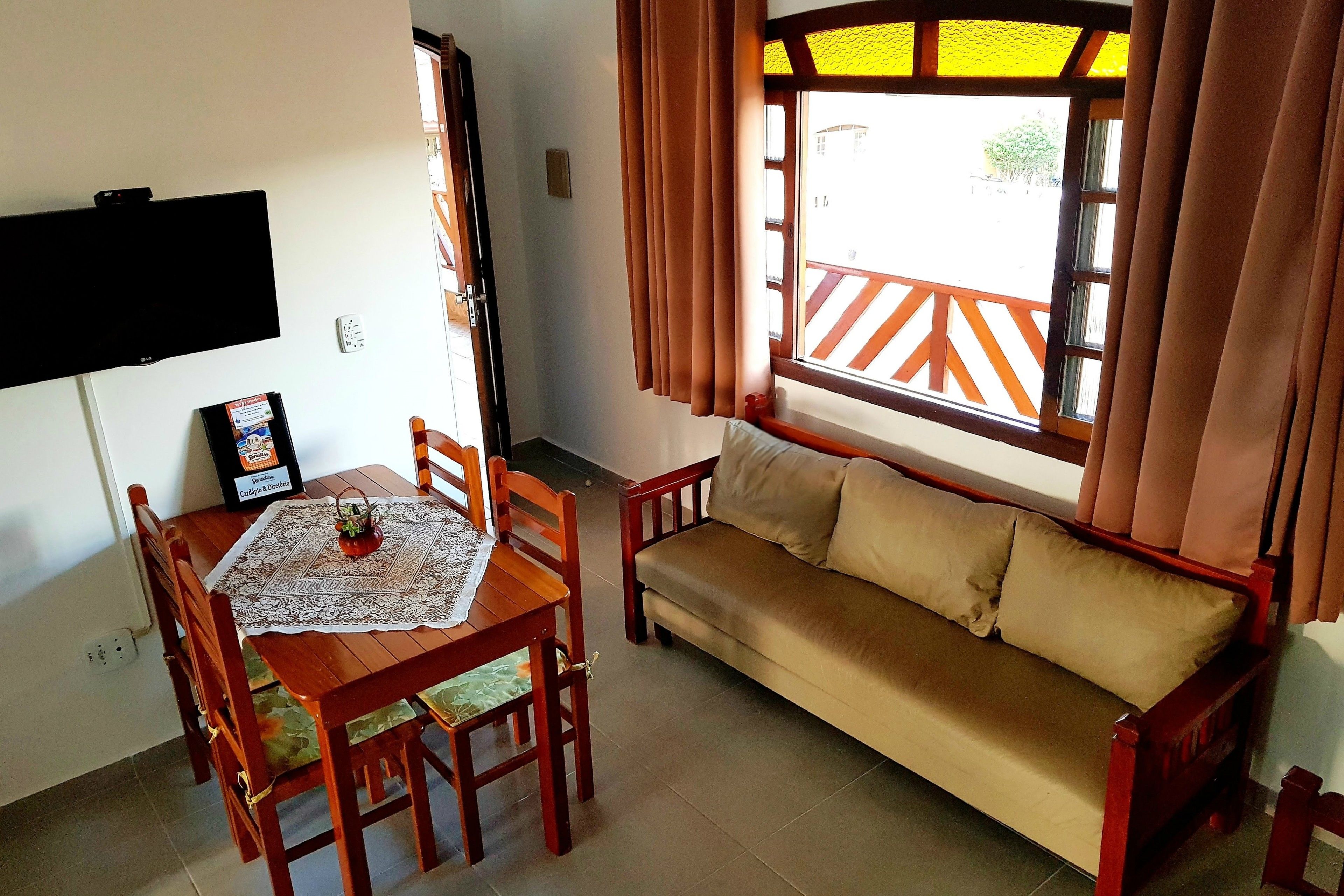 A dining area with a table and chairs, a sofa, a flat-screen TV mounted on the wall, and a window with curtains.