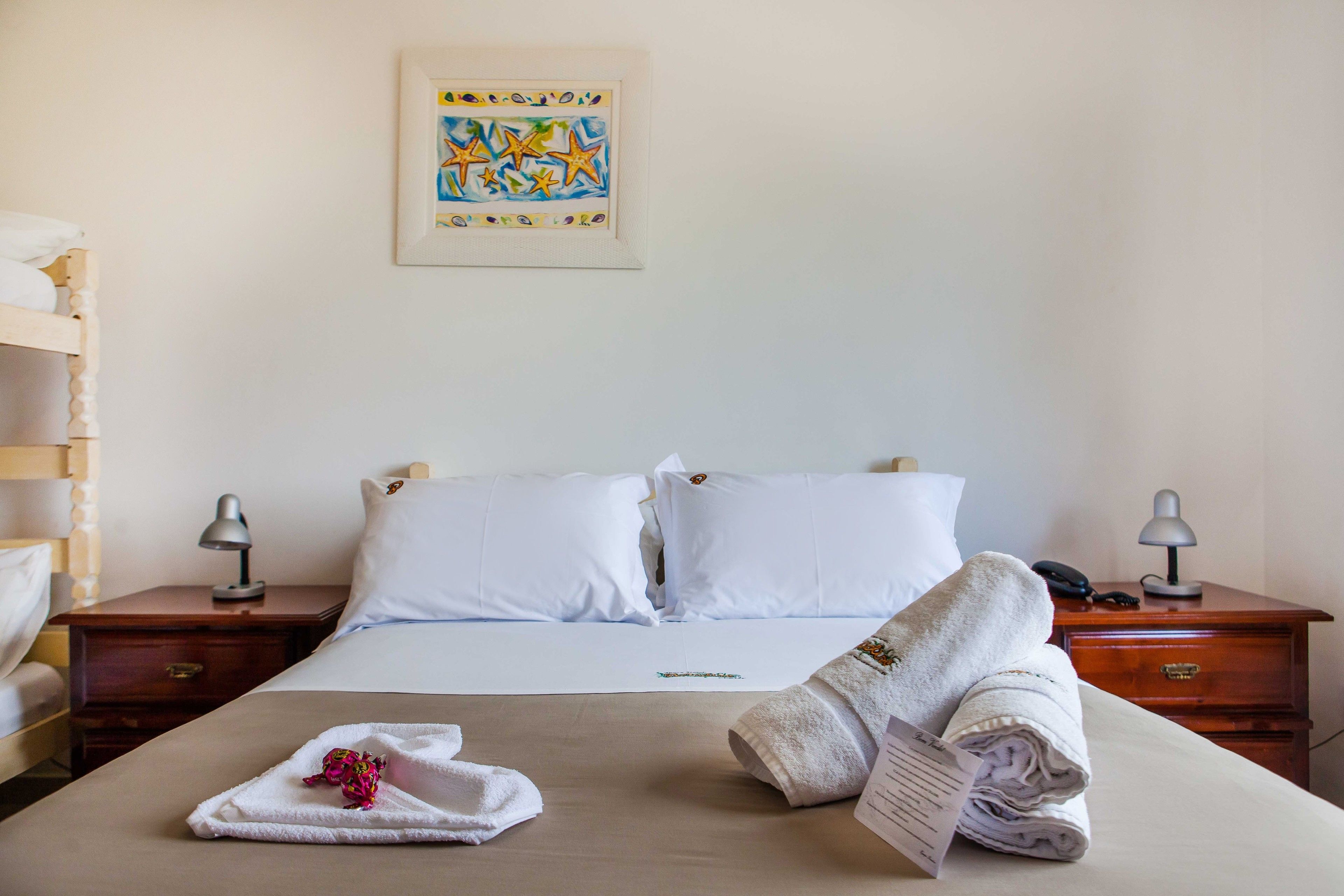 A hotel room with a bed, two bedside tables, a nightstand, a lamp, a framed picture on the wall, and a bunk bed in the background.