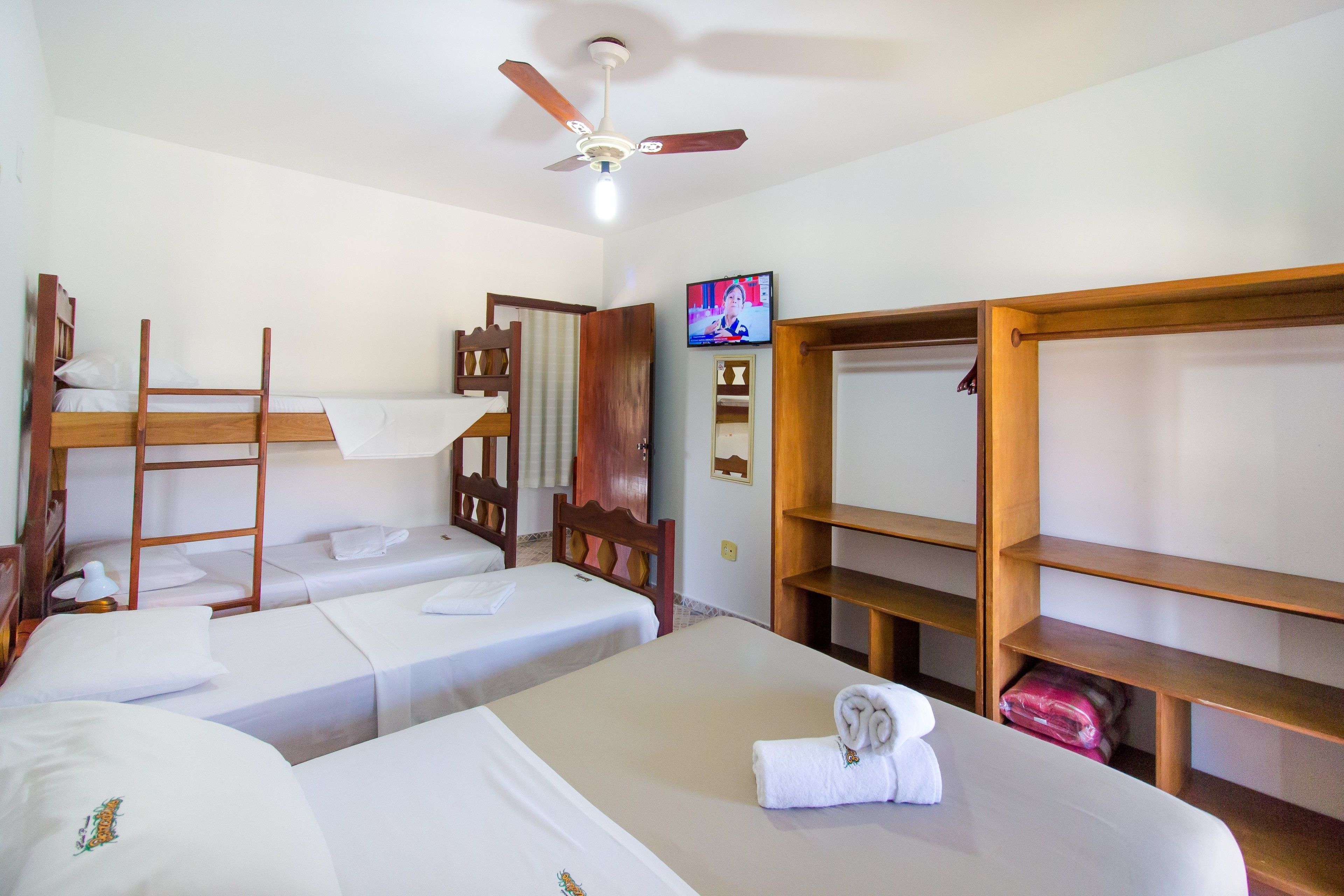 A room with two single beds, a bunk bed, a ceiling fan, a TV, and wooden shelves.