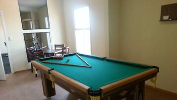 A pool table with a green felt surface, wooden edges, and yellow tassels. There is a dining table with chairs in the background.