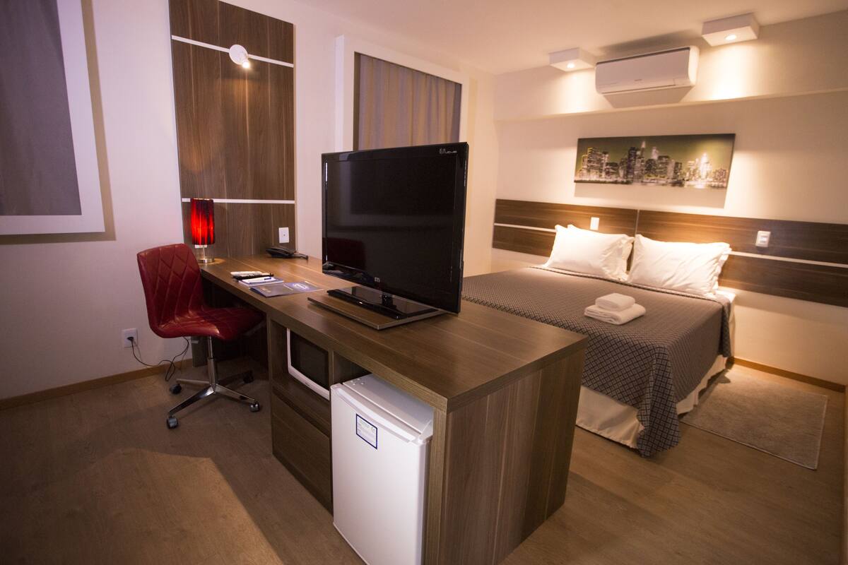 Une chambre d’hôtel avec un bureau en bois, un téléviseur à écran plat, une chaise rouge, un lit orné d’un tableau représentant un paysage urbain et un mini-réfrigérateur blanc.