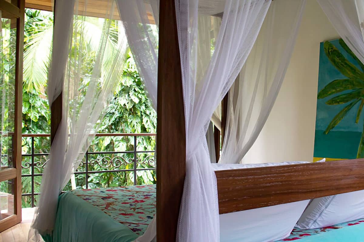 A bed with a wooden headboard and white curtains, a painting of a palm tree, and a balcony with a view of greenery.