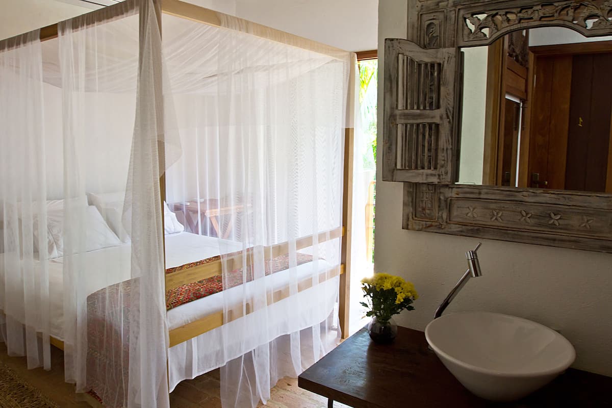 A bedroom with a four-poster bed, a wooden nightstand, a vase with flowers, and a large mirror above a sink.