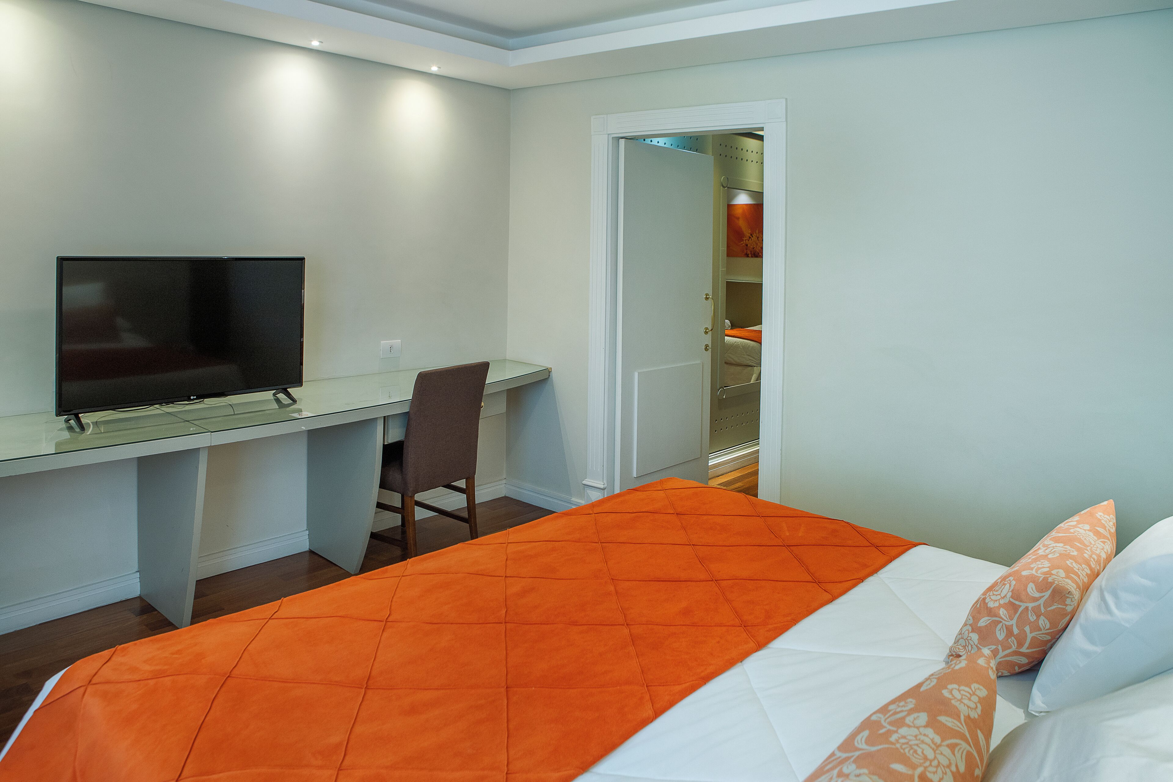 Ein Hotelzimmer mit einem Bett, einer orangefarbenen Tagesdecke, einem weißen Kopfkissen, einem Fernseher auf einem Schreibtisch und einem Stuhl.