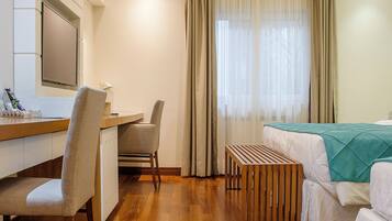 Quarto de hotel com piso de madeira, uma escrivaninha com cadeira, uma cama com manta verde e uma janela com cortinas.