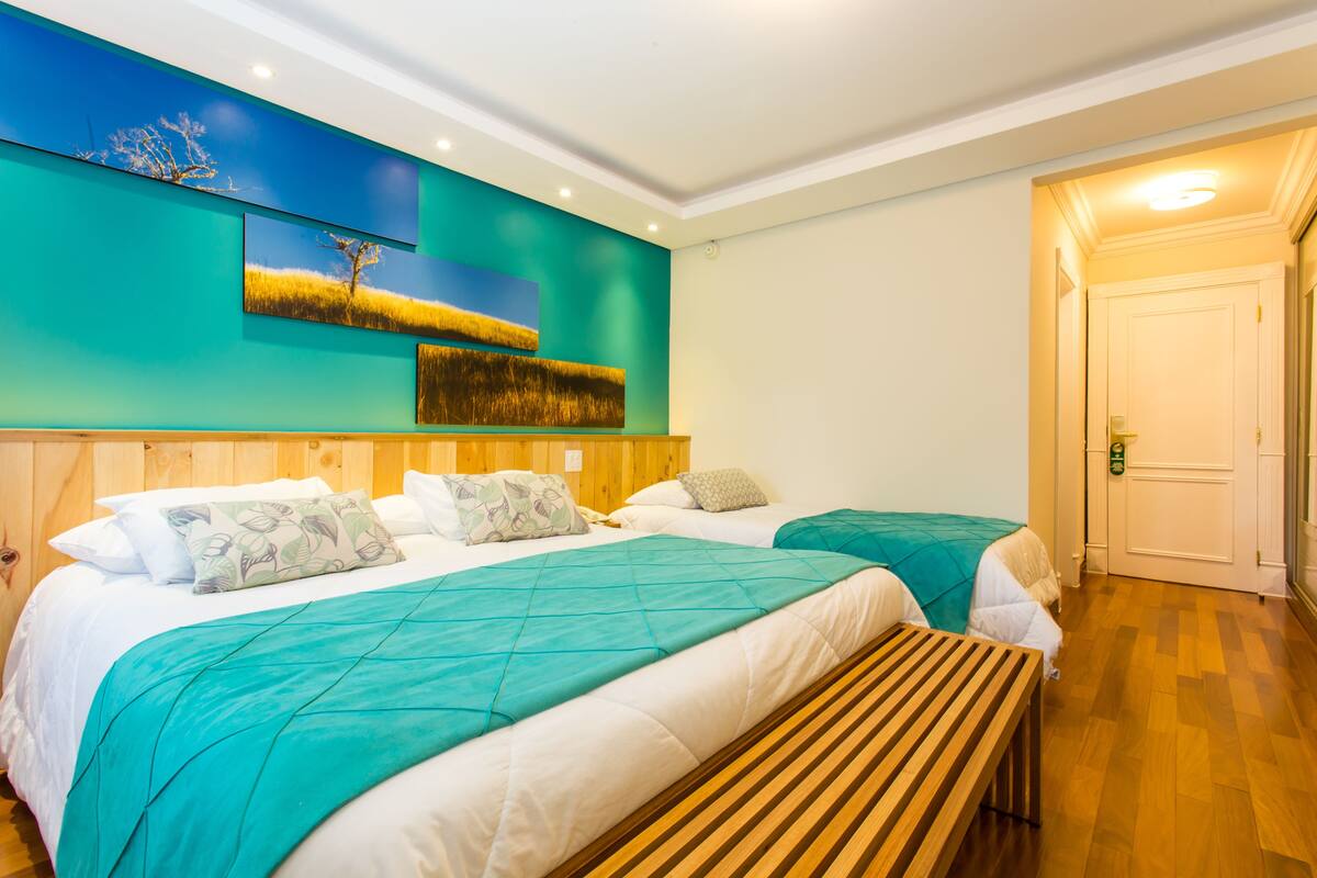 Ein Hotelzimmer mit zwei Betten, einer Holzbänkl und einer türkis farbenen Wand sowie einer Tür.