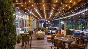 Un patio interior con suelo de madera, mesas y sillas redondas. Hay plantas en macetas y un ventanal que ofrece vistas al exterior.