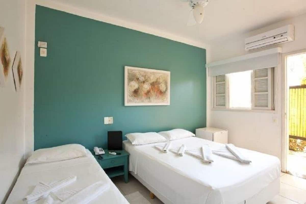 Quarto de hotel com duas camas, uma parede de destaque em tom de verde-água, um quadro, uma escrivaninha com telefone e uma janela com persianas.
