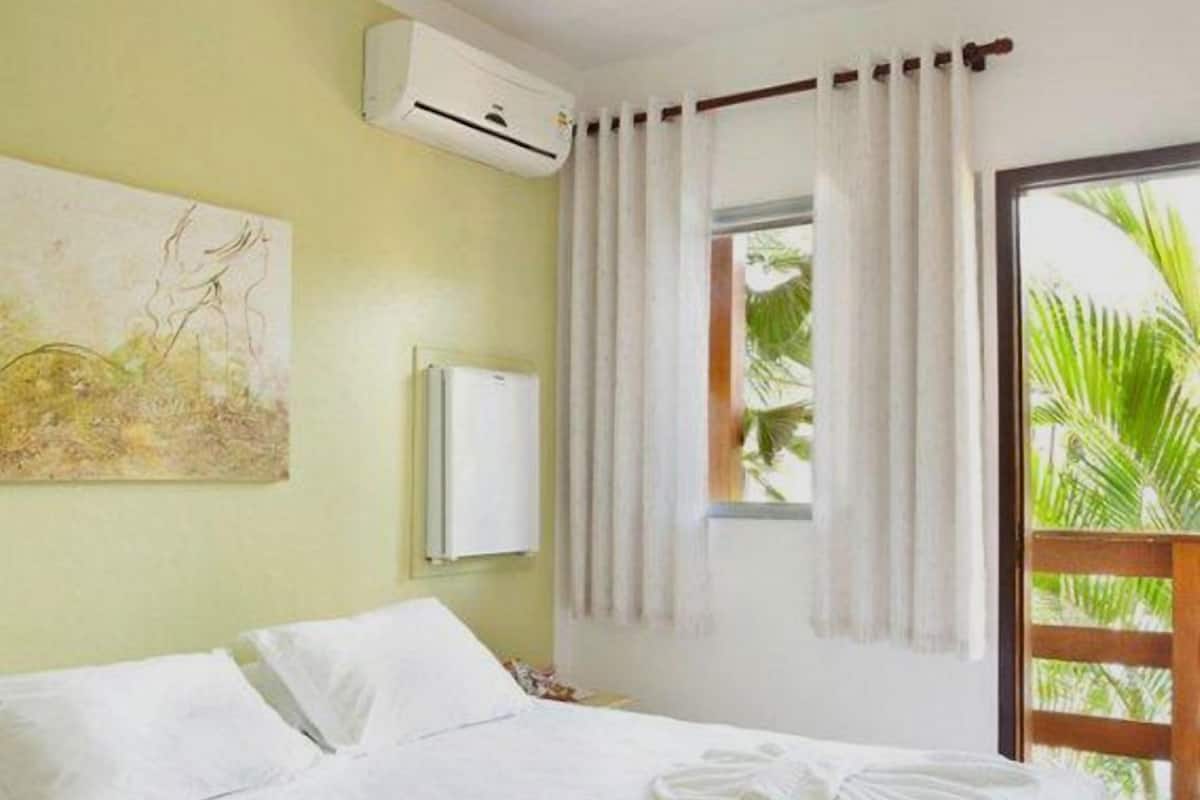 Quarto de hotel com cama, ar condicionado, janela com cortinas e vista para palmeiras.