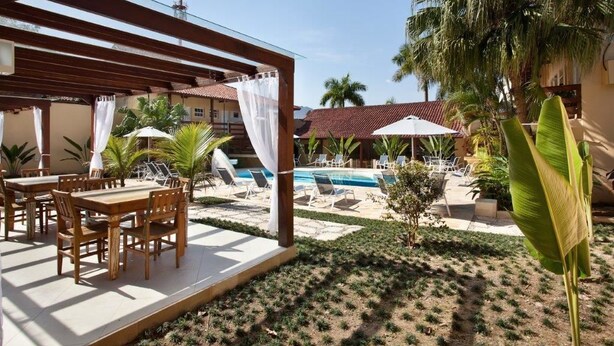 A covered outdoor dining area with wooden tables and chairs, a pool, and palm trees. - Pousada Camburioca (São Sebastião)