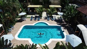 A rectangular pool with a person swimming, surrounded by lounge chairs and umbrellas, with palm trees and a stone wall in the background. - Pousada Camburioca (São Sebastião)