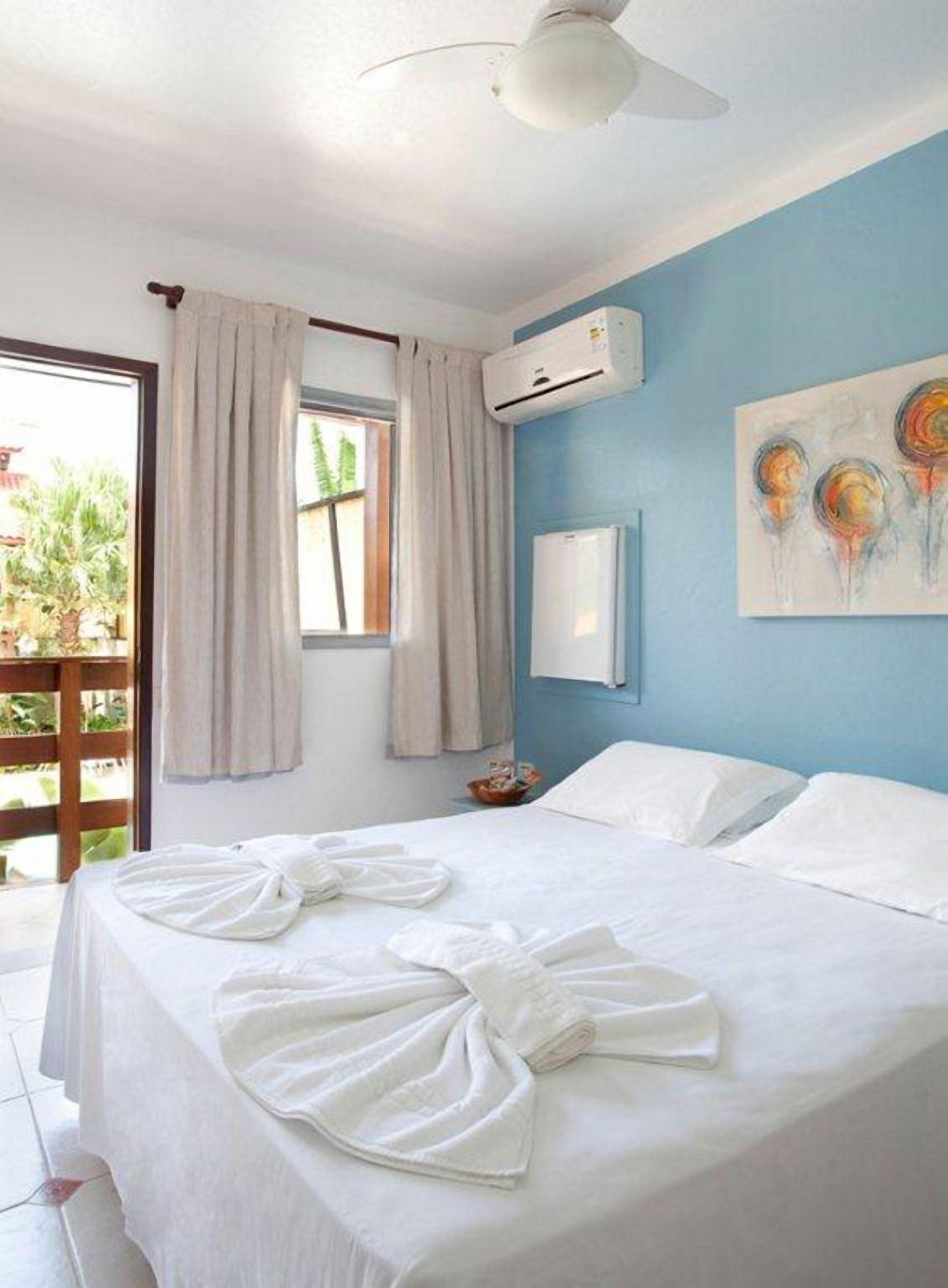 A hotel room with a bed, a ceiling fan, an air conditioning unit, a window with curtains, and a painting on the wall.