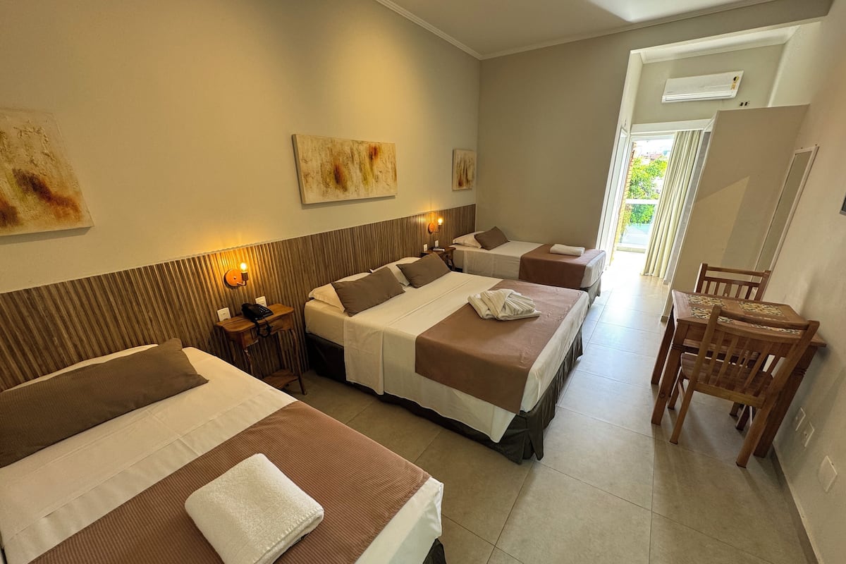 Une chambre d’hôtel avec trois lits, une tête de lit en bois, une petite table avec une lampe et une vue sur l’extérieur.