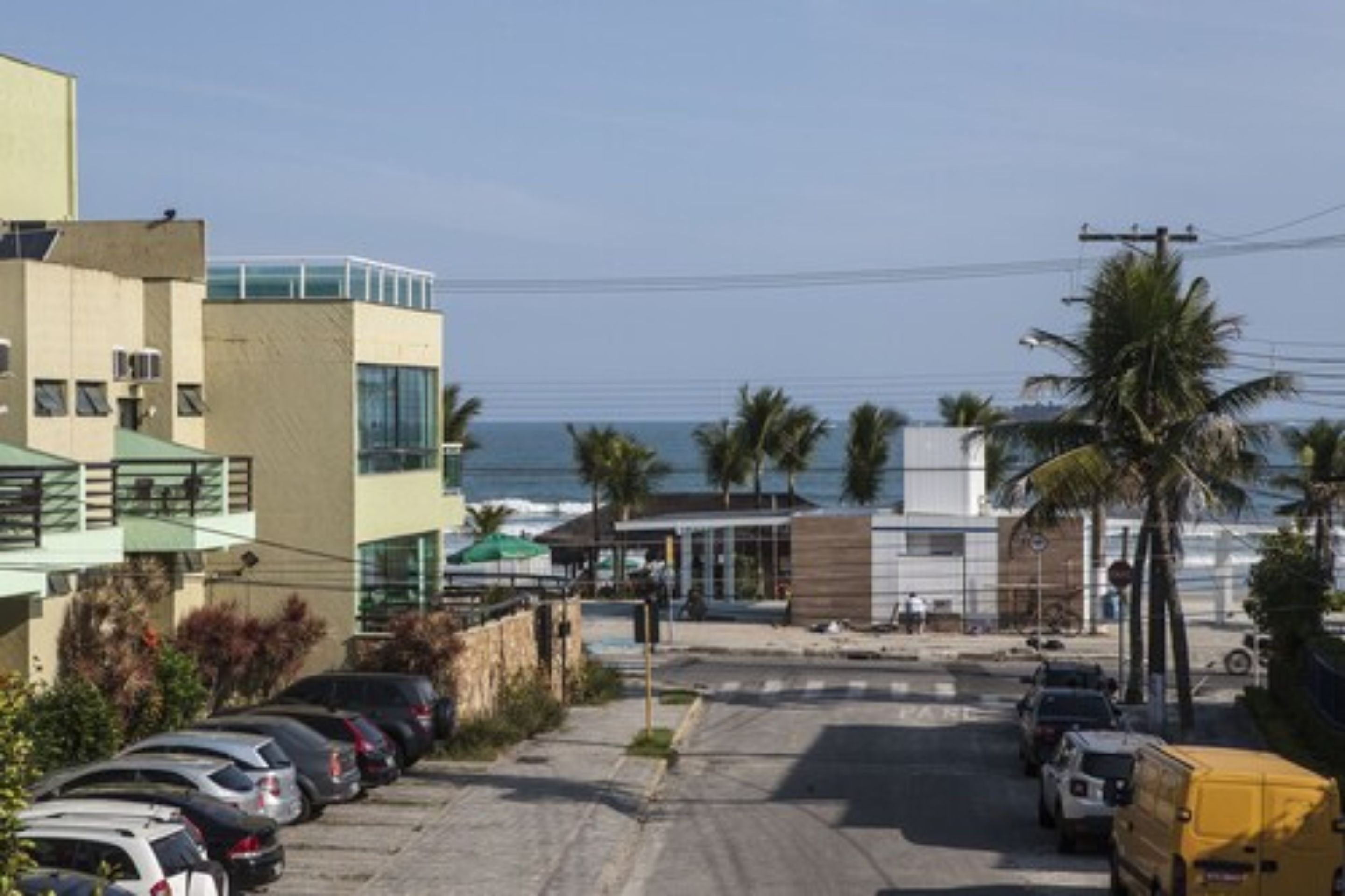 A parking lot with cars and a yellow van, a modern building with large windows, and palm trees along the beachfront.