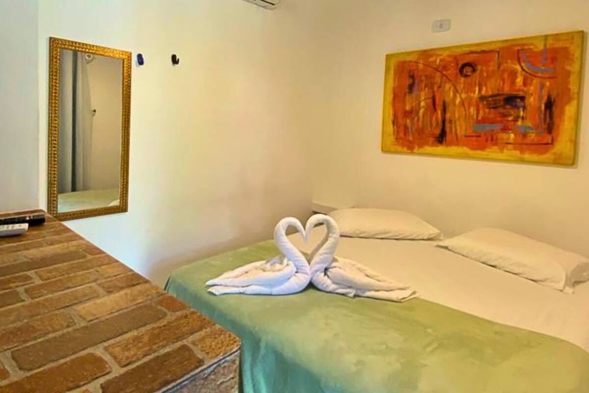 Ein Hotelzimmer mit einem Tisch mit Ziegelmuster, einem Bett mit grüner Bettdecke, zwei zu Schwänen geformten weißen Handtüchern, einem großen Spiegel und einem Gemälde an der Wand.