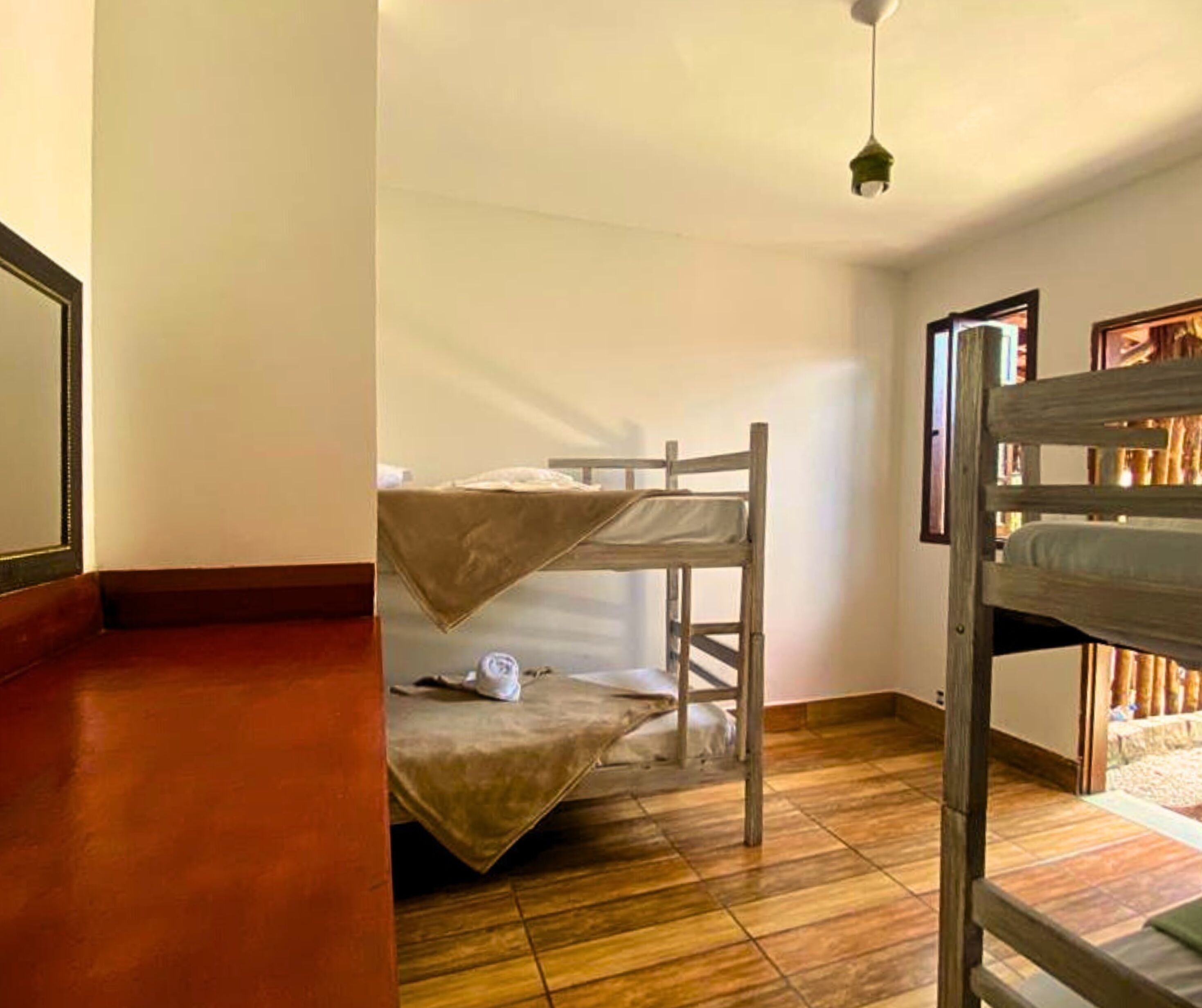 A room with two bunk beds, wooden flooring, a ceiling light, and a window with a view of the outside.