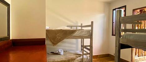 A room with two bunk beds, wooden flooring, a ceiling light, and a window with a view of the outside.