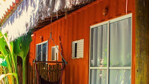 A room with a wooden exterior, a large window with curtains, a hammock, and a thatched roof. - Pousada Papaya (Ubatuba)