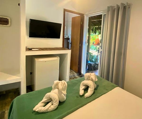 A hotel room with a bed, a television on a stand, a small refrigerator, and a view of the outside through a door.