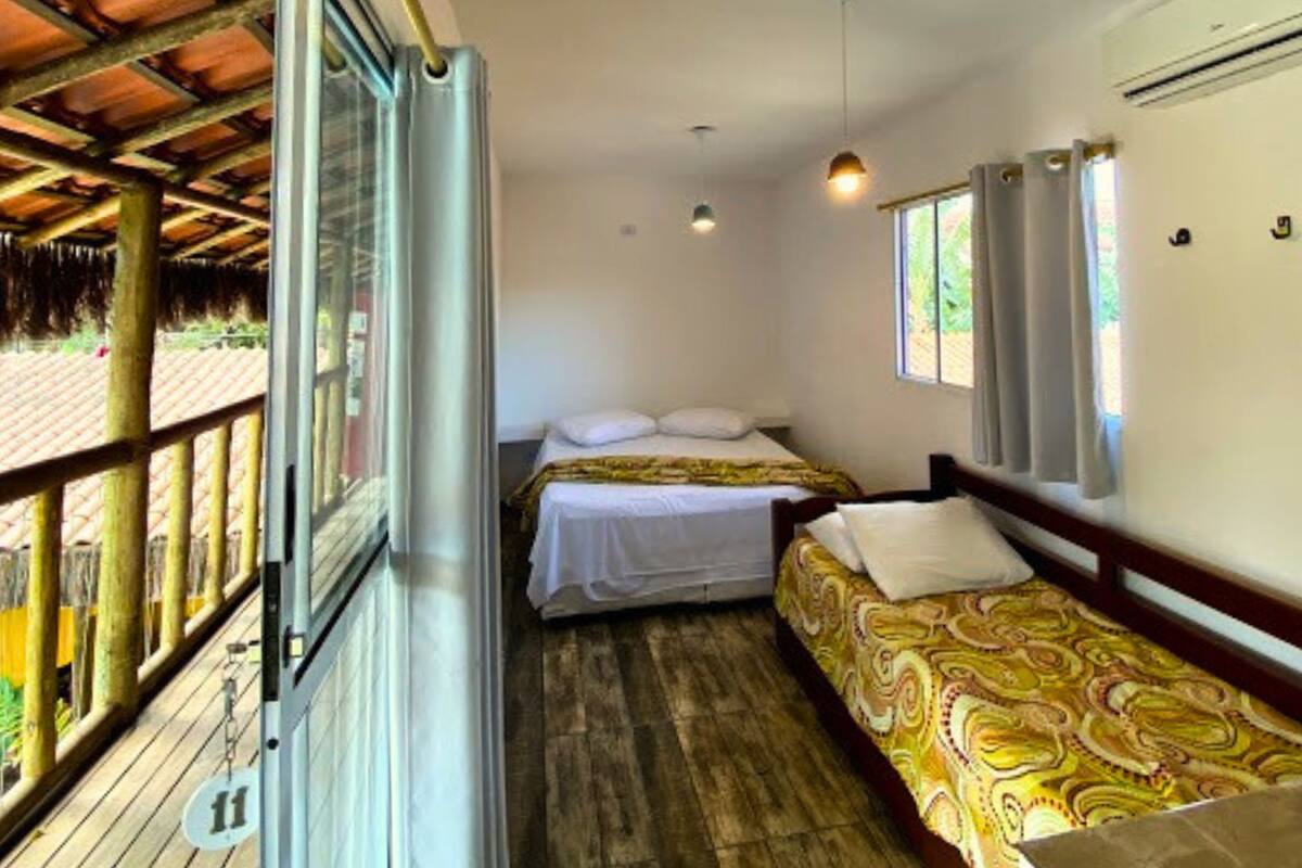 Ein Zimmer mit zwei Betten, einem fensterbelaubten Fenster, einem Deckenventilator und einem Balkon mit Holzgeländer.