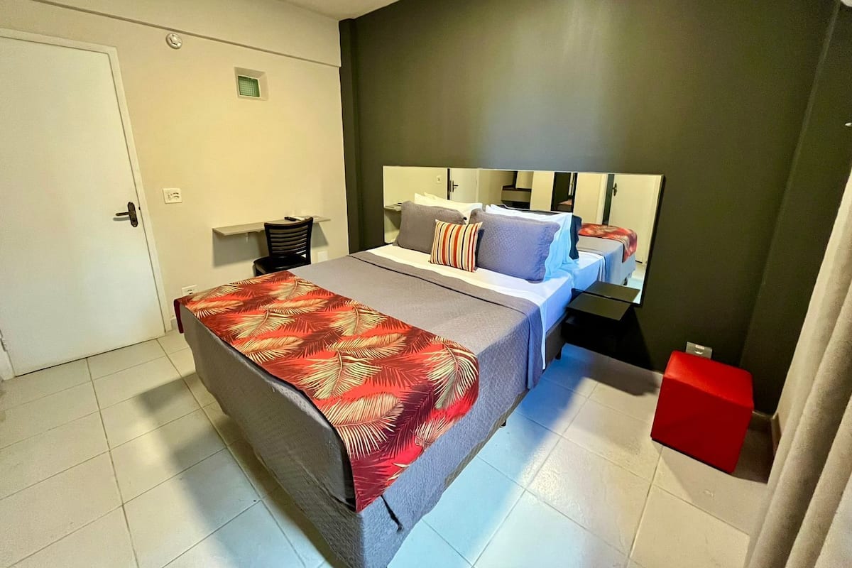 A hotel room with a bed, a desk, a chair, a mirror, and a red object on the floor.