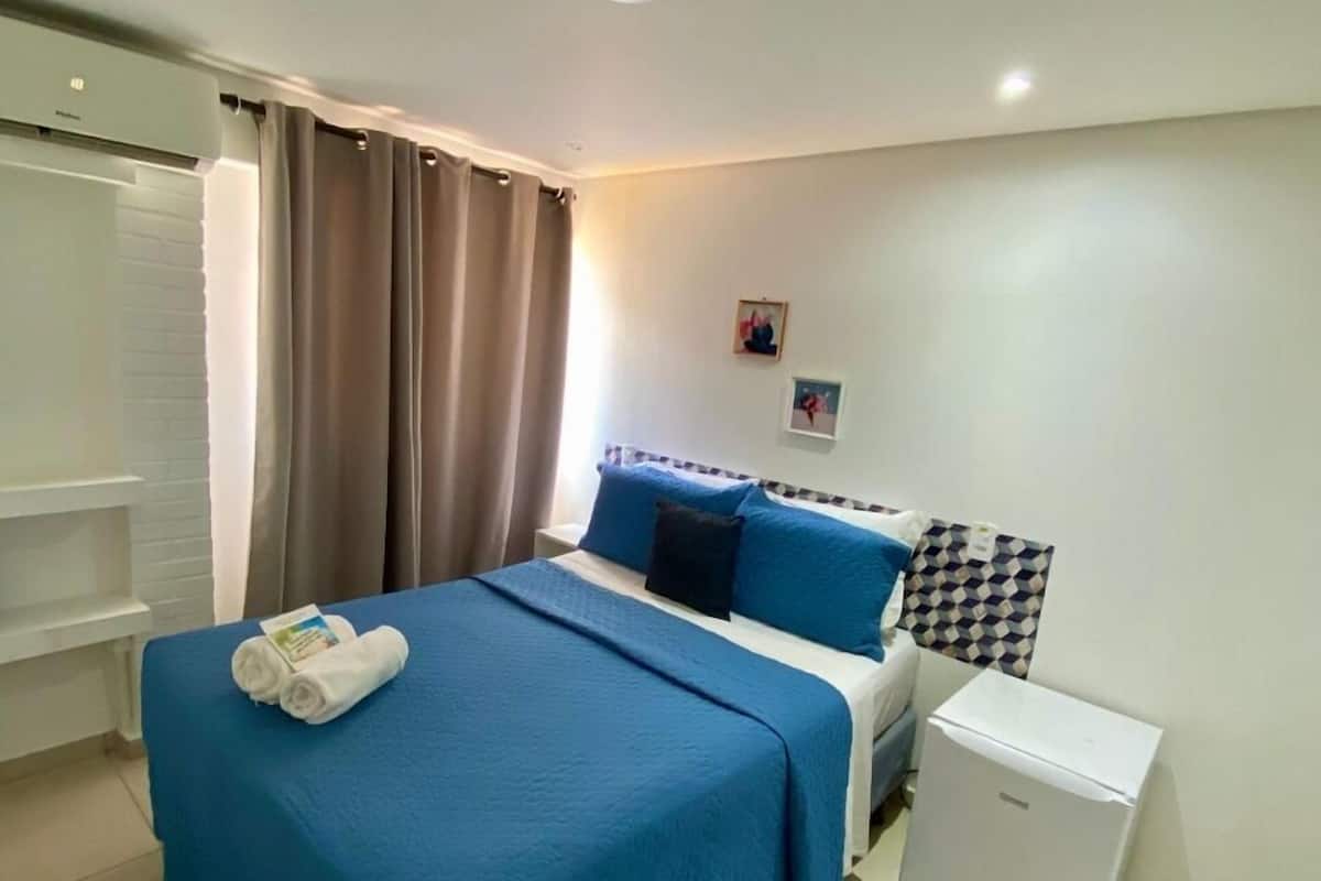 A hotel room with a bed, a nightstand, a small refrigerator, a window with curtains, and a wall-mounted air conditioner.