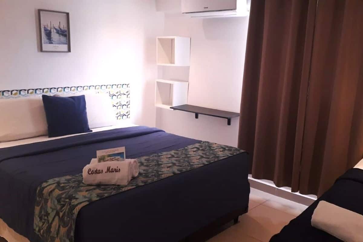 A hotel room with a bed, a side table, a small shelf, a window with curtains, and a wall-mounted air conditioner.