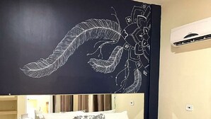 Une chambre d’hôtel avec un lit, une table de chevet, un mur orné d’un motif de plumes et de fleurs, et un livre posé sur le lit.