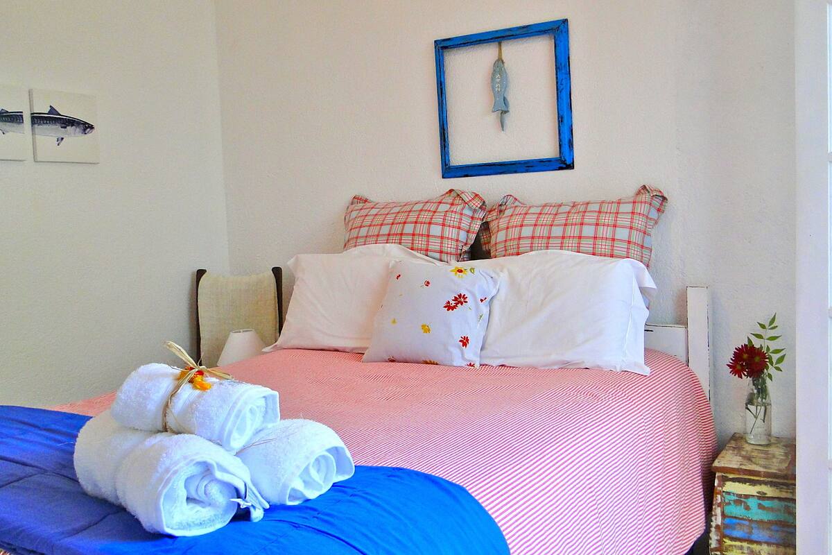 A neatly made bed with a pink and white checkered bedspread, white pillows with floral patterns, and a blue blanket. There are rolled white towels on the bed, and a framed fish decoration on the wall.