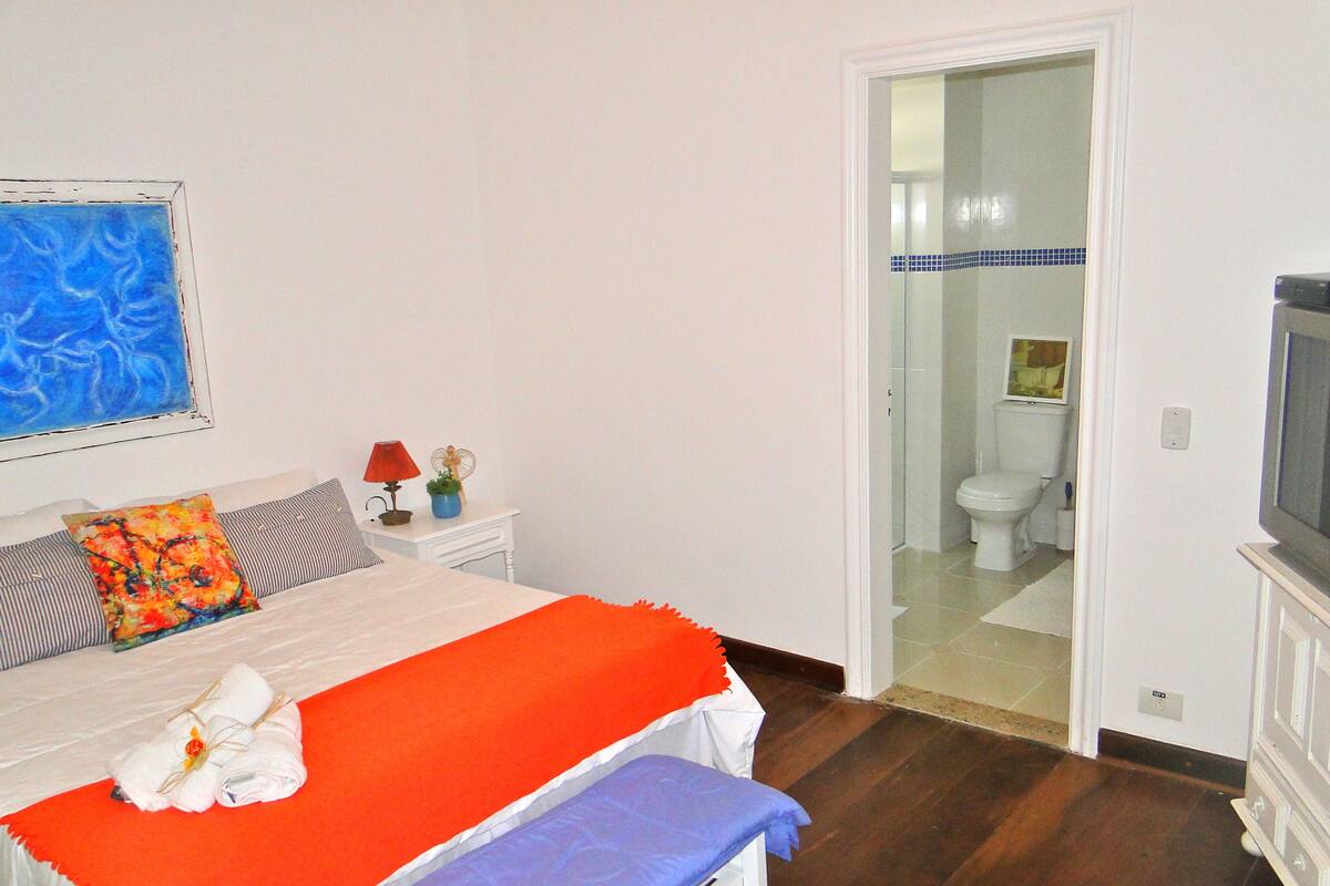 A bedroom with a bed, a bedside table with a lamp, a TV, and a bathroom visible through an open door.