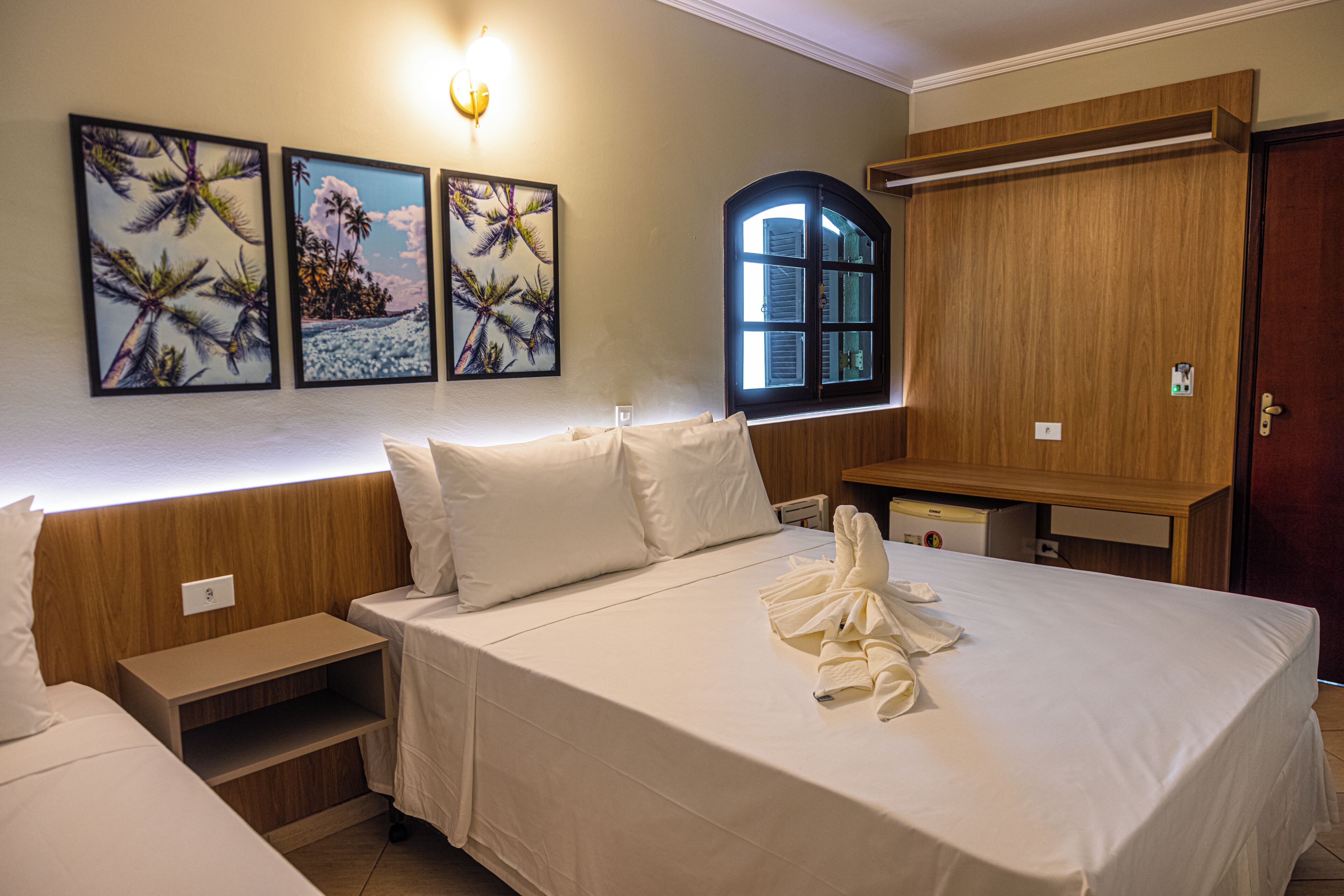 A hotel room with a large bed, a wooden wardrobe, a small desk, and a window with a view of palm trees.