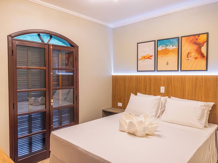 Quarto de hotel com cama, cabeceira de madeira, mesa de cabeceira, guarda-roupa de madeira e três quadros na parede.