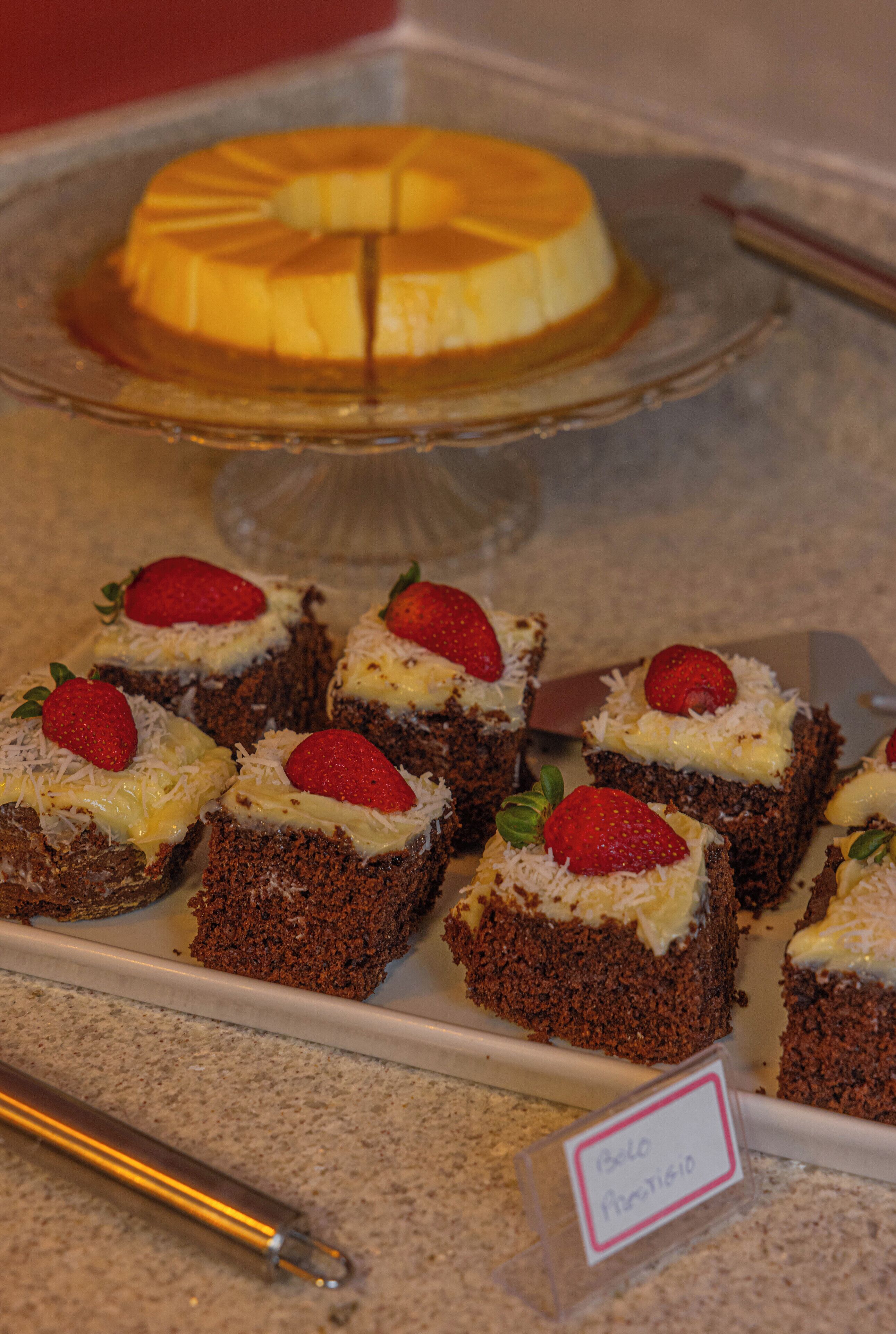 A dessert display with a flan and several chocolate cakes topped with strawberries and coconut.