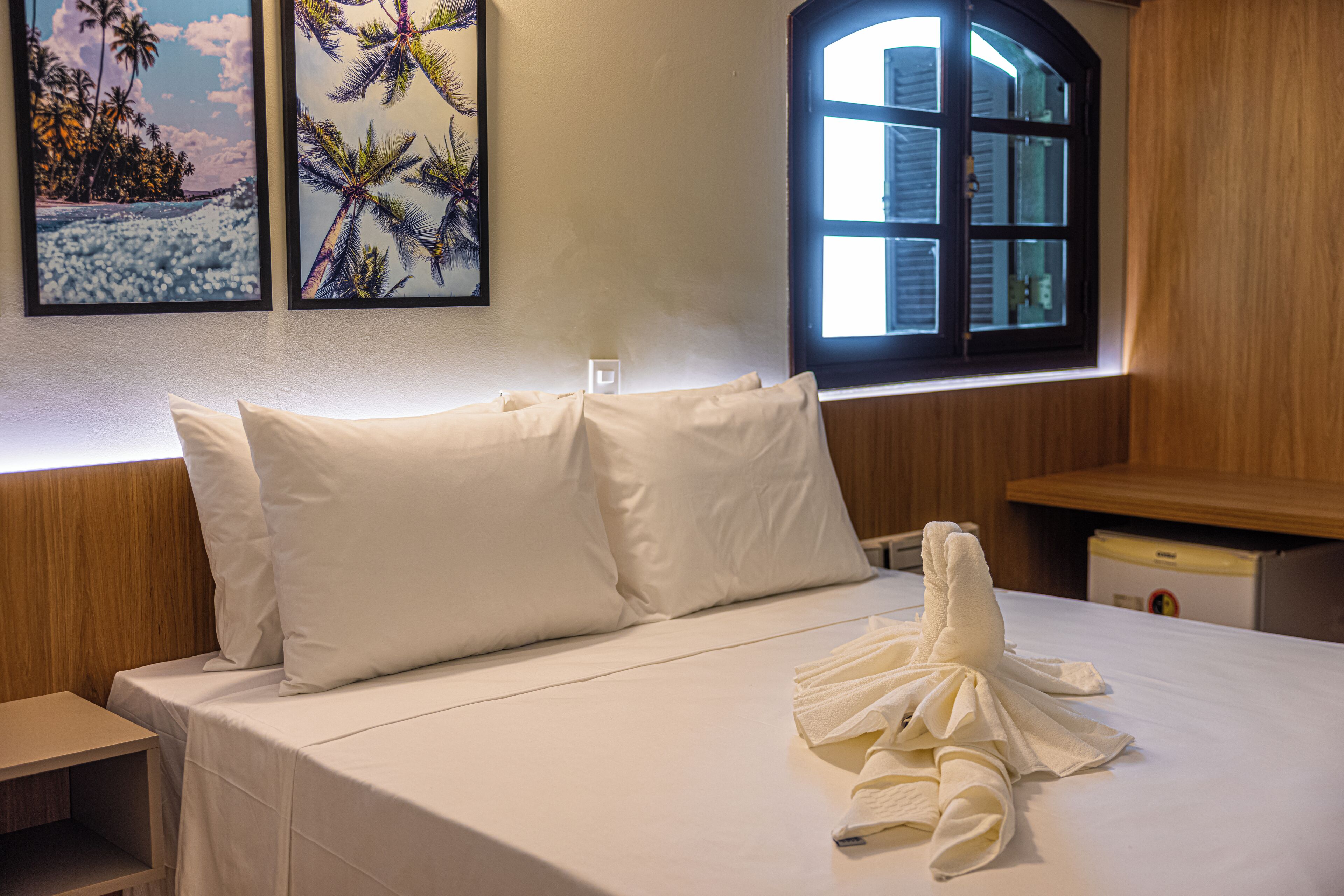 A neatly made bed with white linens and pillows, a towel on the bed, and a window with a view of palm trees.