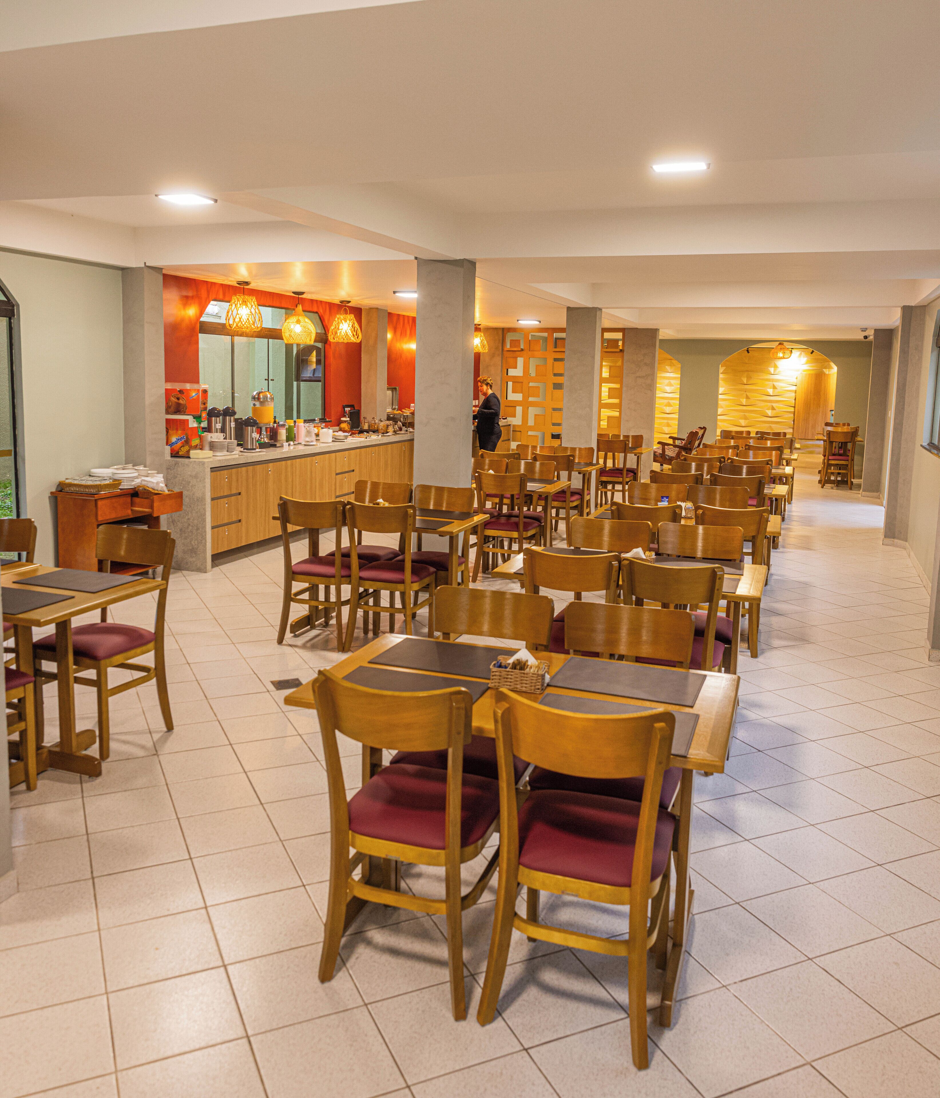 A hotel dining area with rows of wooden tables and chairs, a counter with food and beverages, and a person working at the counter.