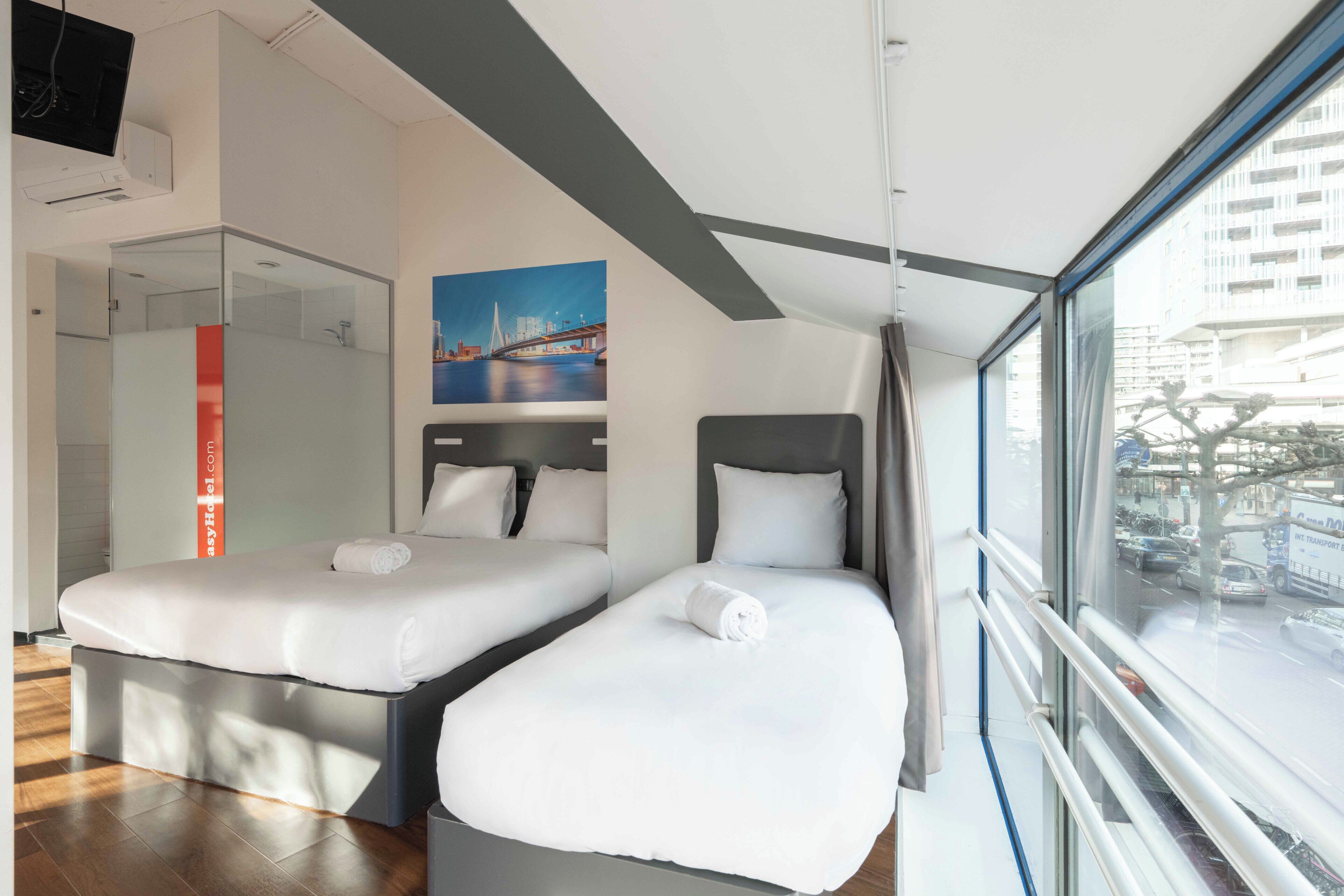 A modern hotel room with two beds, a large window with a city view, and a painting on the wall.