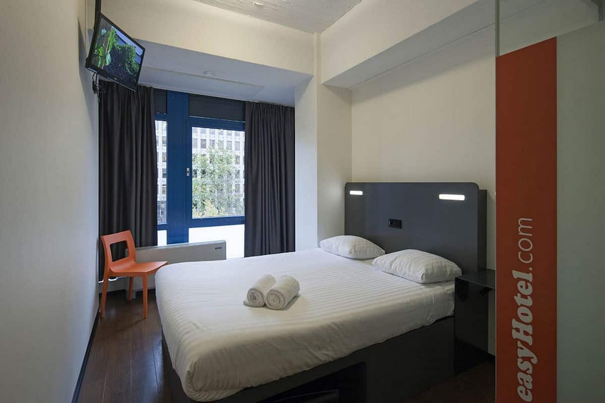 A hotel room with a bed, a TV mounted on the wall, a chair, and a window with curtains.
