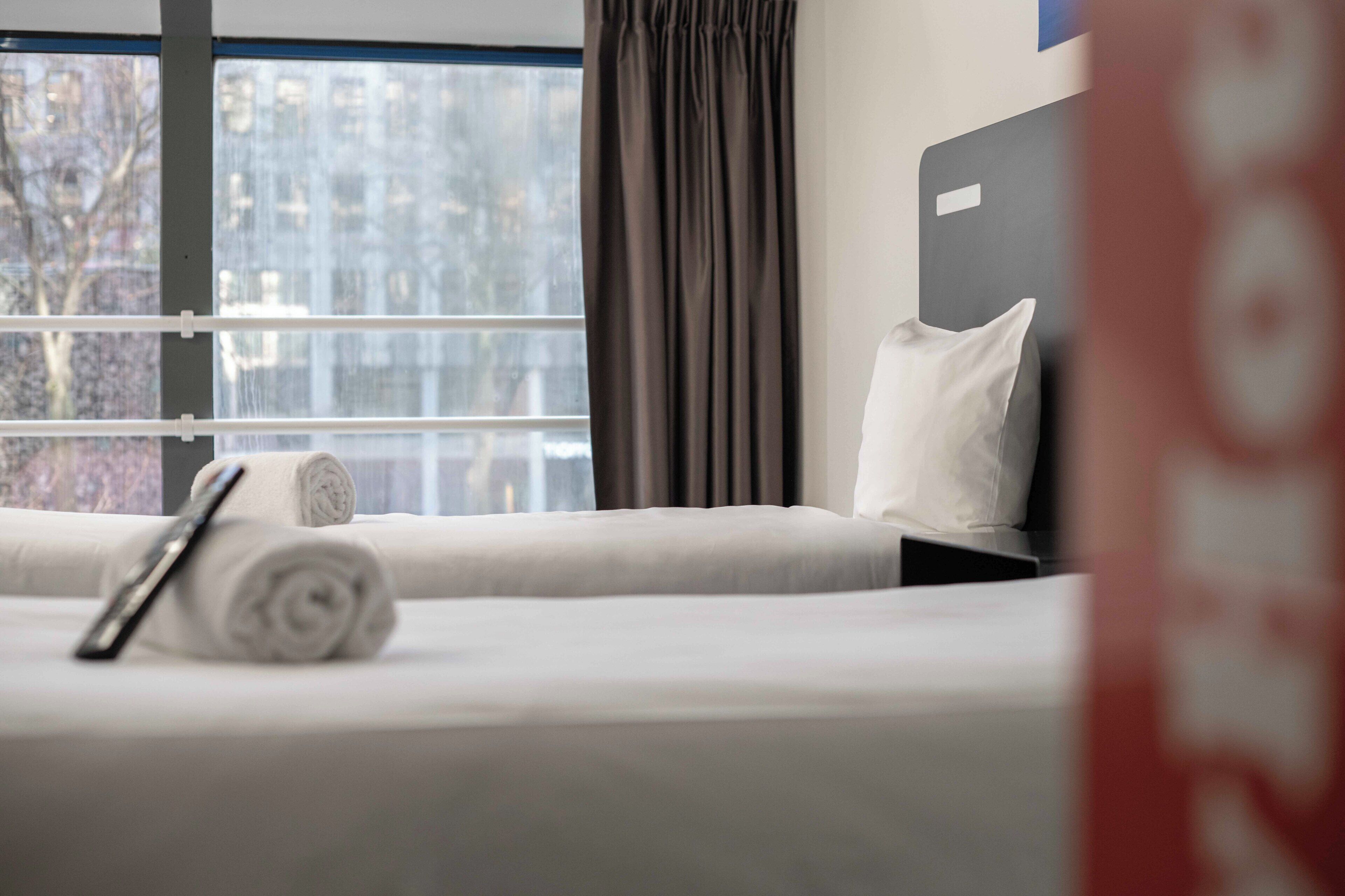Una camera d'albergo con un letto, un asciugamano arrotolato e una finestra con tende.