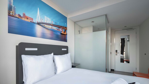 A hotel room with a bed, white linens, a painting of a cityscape, a mirror, and a door leading to a bathroom.