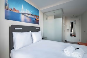 A hotel room with a bed, white linens, a painting of a cityscape, a mirror, and a door leading to a bathroom.