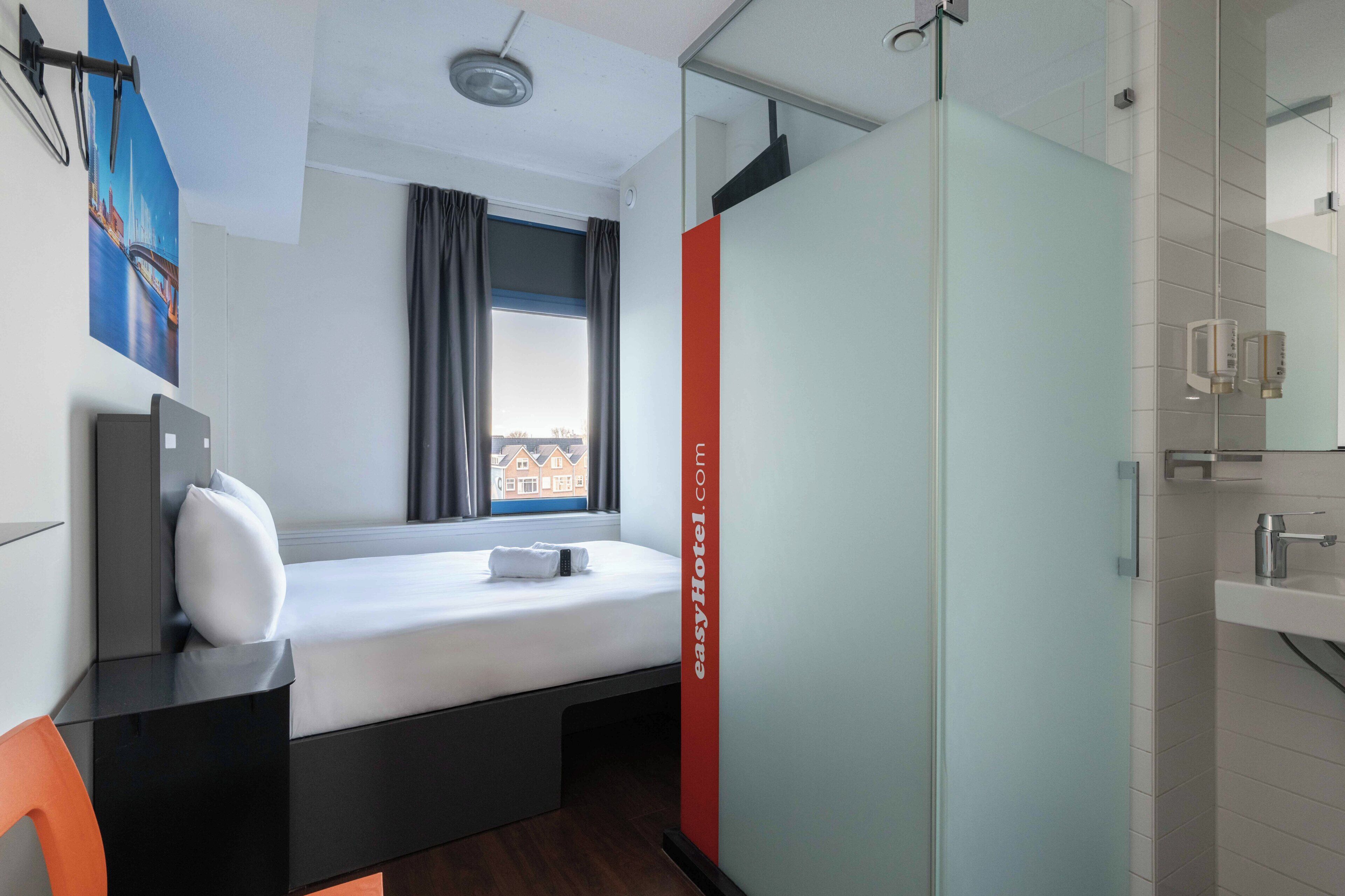 A hotel room with a bed, a window with curtains, a shower area, and a wall-mounted mirror.