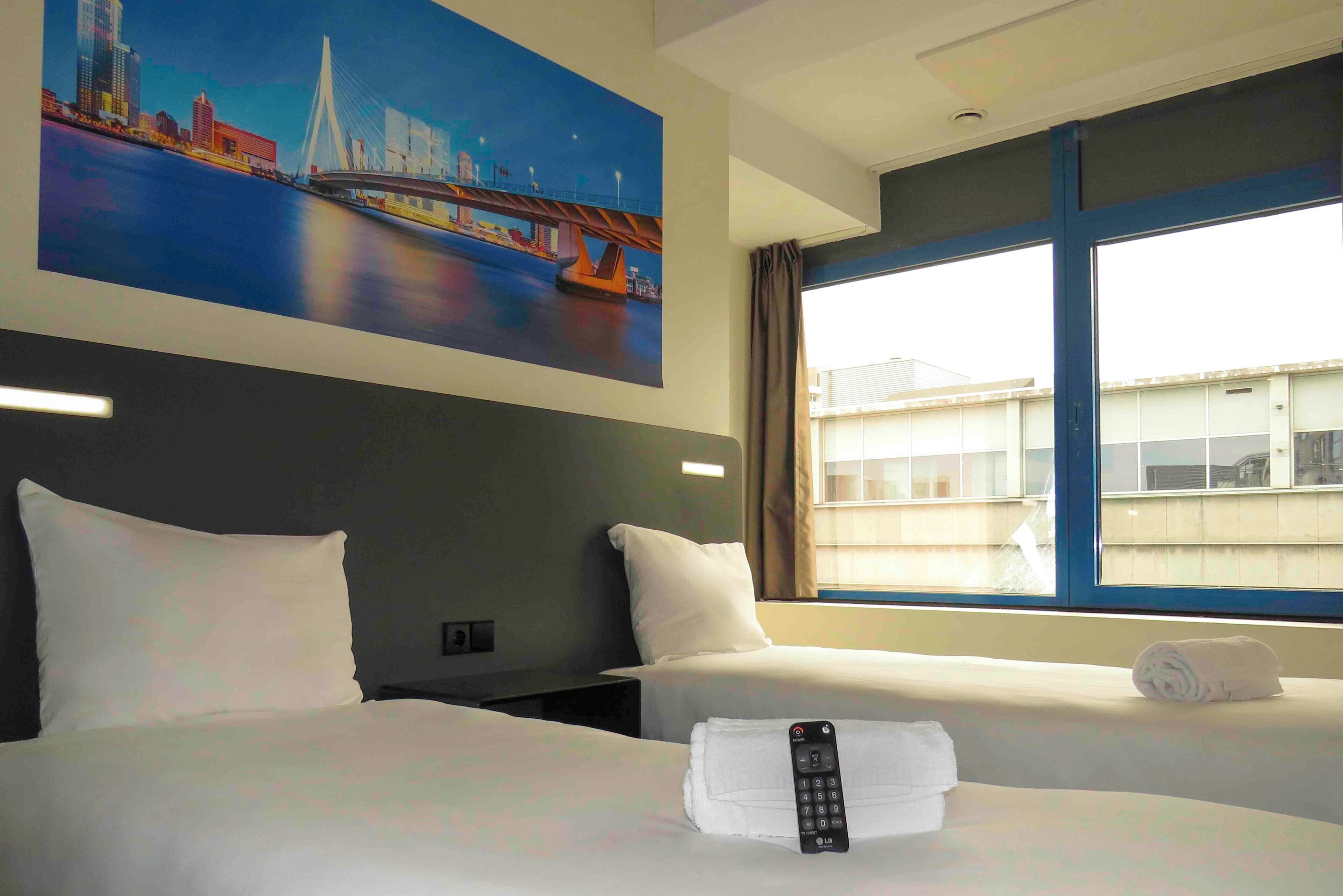 A hotel room with two beds, a painting of a cityscape, a window with curtains, and a remote control on the bed.