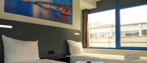 Habitación de hotel con dos camas, un cuadro de un paisaje urbano, una ventana con cortinas y un control remoto sobre la cama.